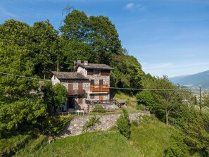 Un bâtiment en pierre doté d’un balcon en bois, entouré d’une végétation luxuriante et offrant une vue dégagée sur les montagnes lointaines.