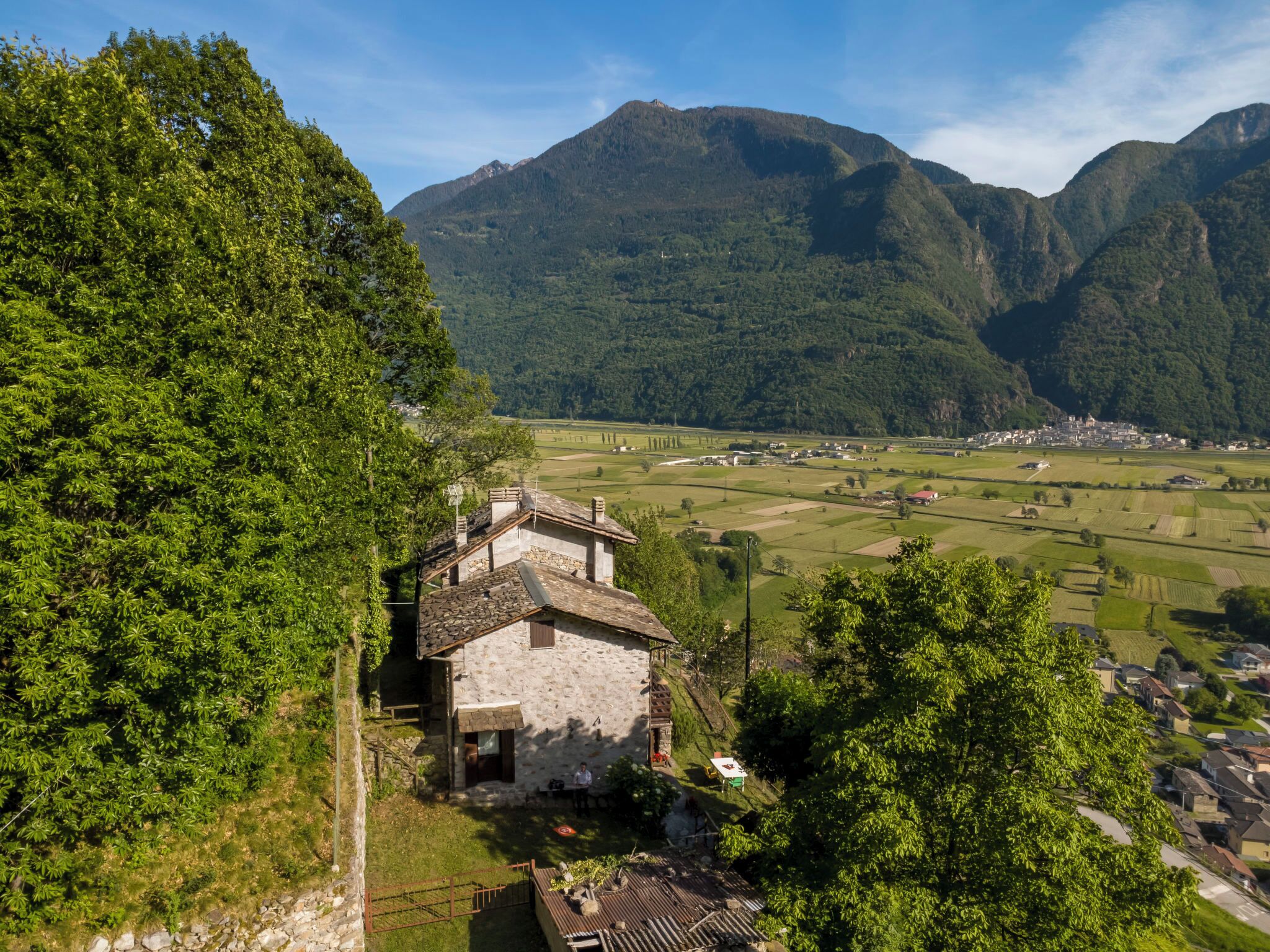 A stone house with a sloped roof, surrounded by lush greenery and a mountainous backdrop.