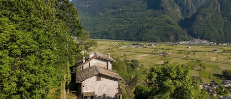 A stone house with a sloped roof, surrounded by lush greenery and a mountainous backdrop.