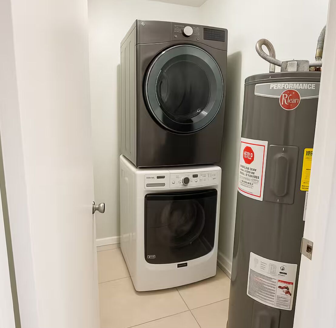 A laundry room with a stacked washer and dryer, a water heater, and a door with a safety sign.