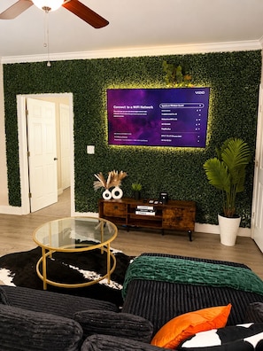 Ein modernes Wohnzimmer mit grüner Wand, einem Flachbildfernseher, einem Glastisch, einer Couch und einer Holzkommode.