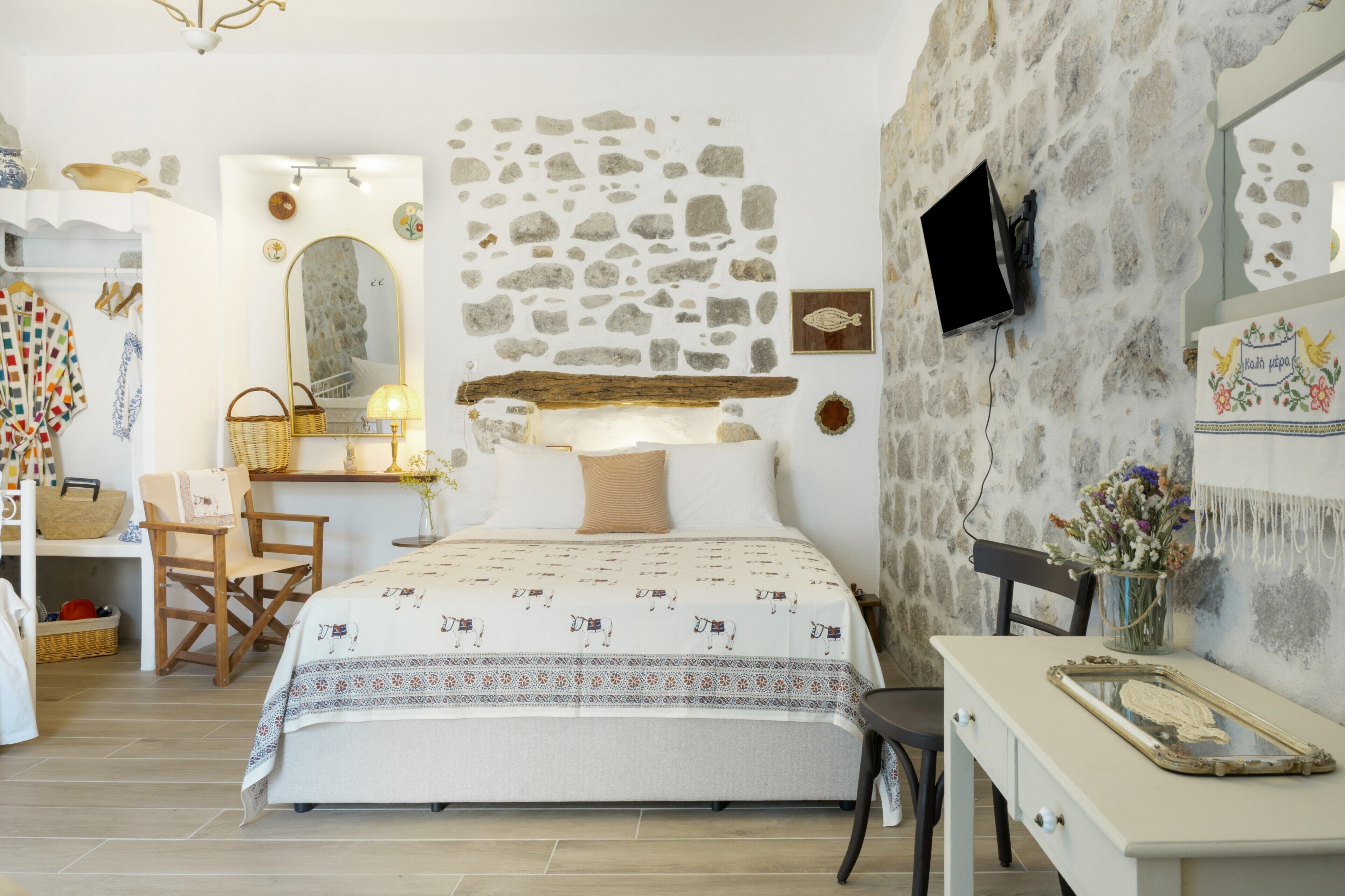 A bedroom with a stone wall, a bed with patterned bedding, a desk with a lamp, and a television mounted on the wall.