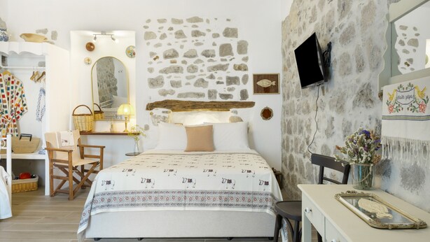 Quarto com parede de pedra, cama com roupa de cama estampada, escrivaninha com abajur e televisão fixada na parede.