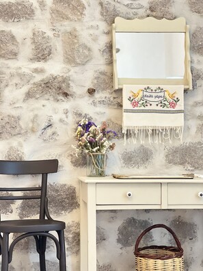 A rustic stone wall, a wooden chair, a white desk with drawers, a woven basket, and a mirror with a decorative cloth.