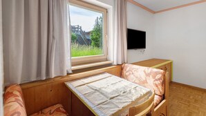 A room with a bed, a TV, a window with curtains, and a view of greenery outside. - Apartment '7 Residence Ciasa Ulli Typ A' with Mountain View, Shared Terrace and Wi-Fi (San Cassiano)