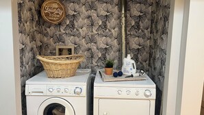 A laundry area with two washing machines, a basket, a potted plant, and a bottle of laundry detergent. - Family Friendly Suite (Langley Township)