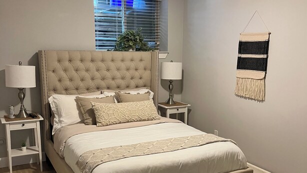 A bedroom with a tufted headboard, a large bed, two bedside tables with lamps, a window with blinds, a wall-mounted macrame piece, and a small plant on the headboard. - Family Friendly Suite (Langley Township)