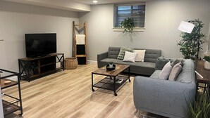 A living room with a grey sofa, a TV on a wooden stand, a wooden ladder, a window with blinds, and a plant. - Family Friendly Suite (Langley Township)
