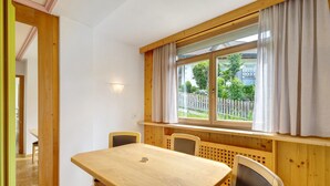A wooden dining table with chairs, a window with curtains, and a view of a fence and greenery outside. - Apartment '6 Residence Ciasa Ulli Typ A' with Mountain View, Shared Terrace and Wi-Fi (San Cassiano)