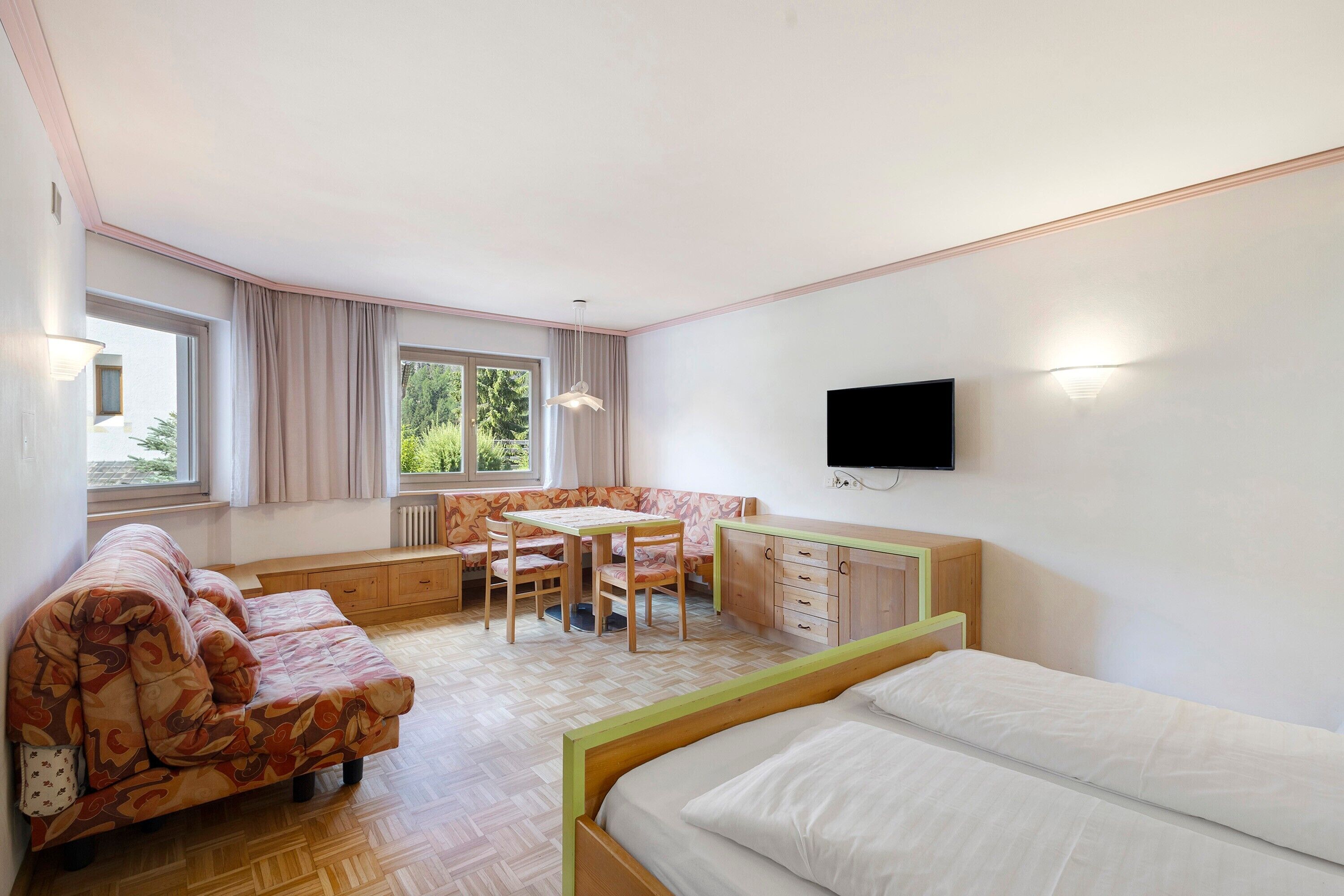 Una habitación con una cama, un sofá, una mesa de comedor con sillas, un televisor montado en la pared y ventanas amplias.