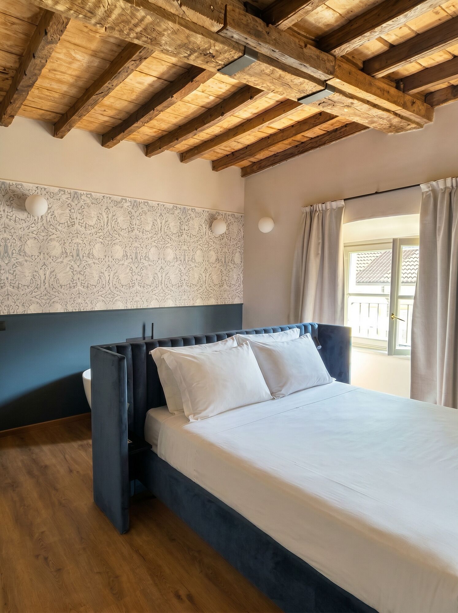 Un dormitorio con techo de madera, pared con diseño, cama con ropa blanca y ventana con cortinas.