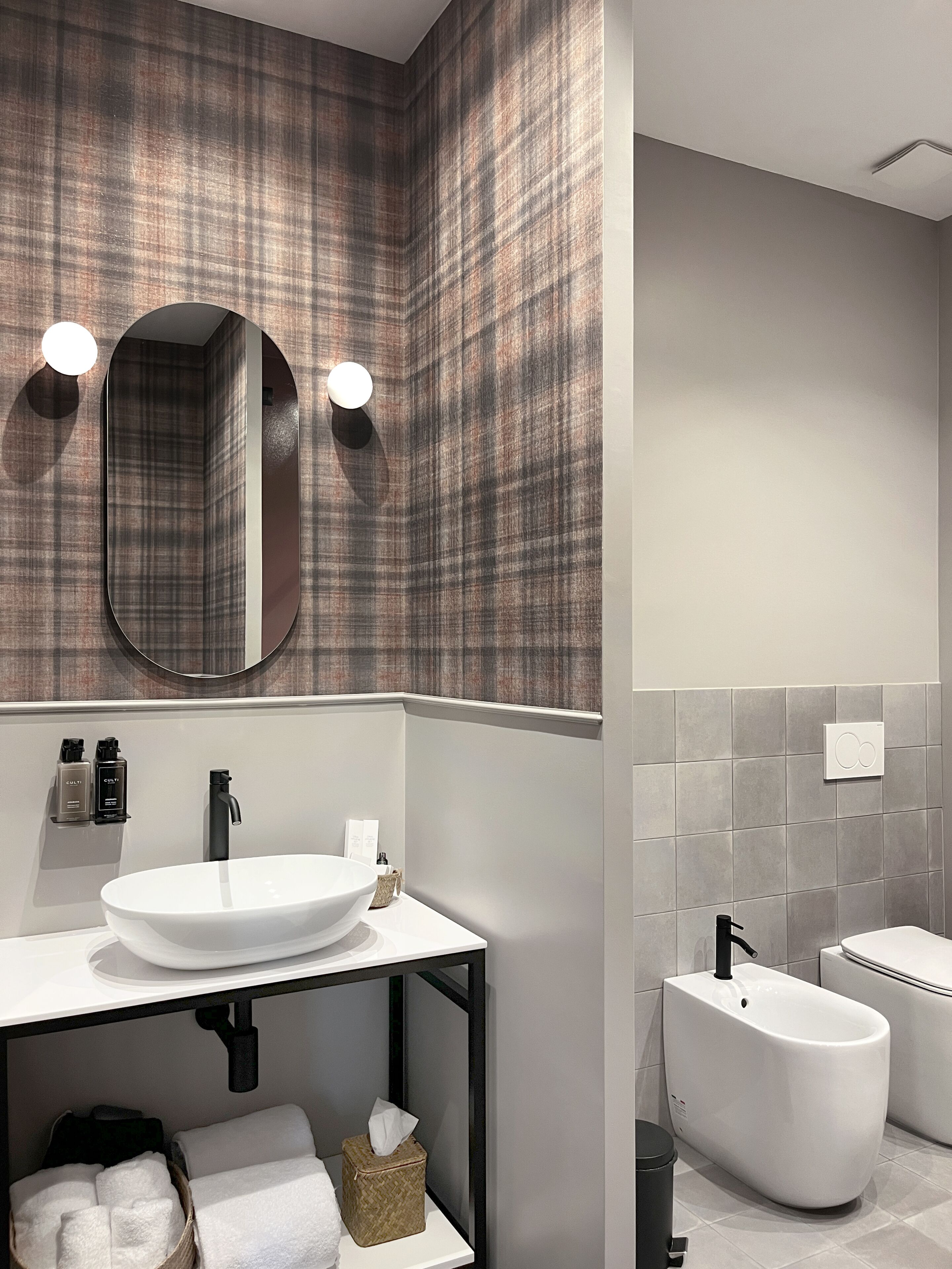Un bagno moderno con uno specchio grande, due luci a parete, un lavandino bianco, un rubinetto nero, un water bianco e un bidet.