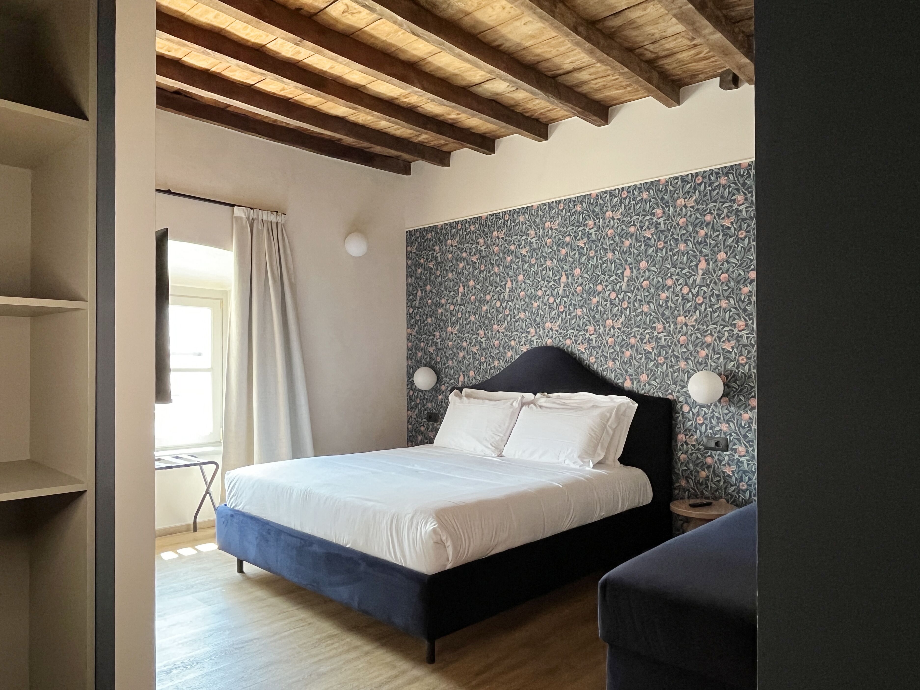 Une chambre à coucher avec un lit, une table de chevet, un mur recouvert d’un papier peint à motifs floraux, un plafond en bois et une fenêtre avec des rideaux.