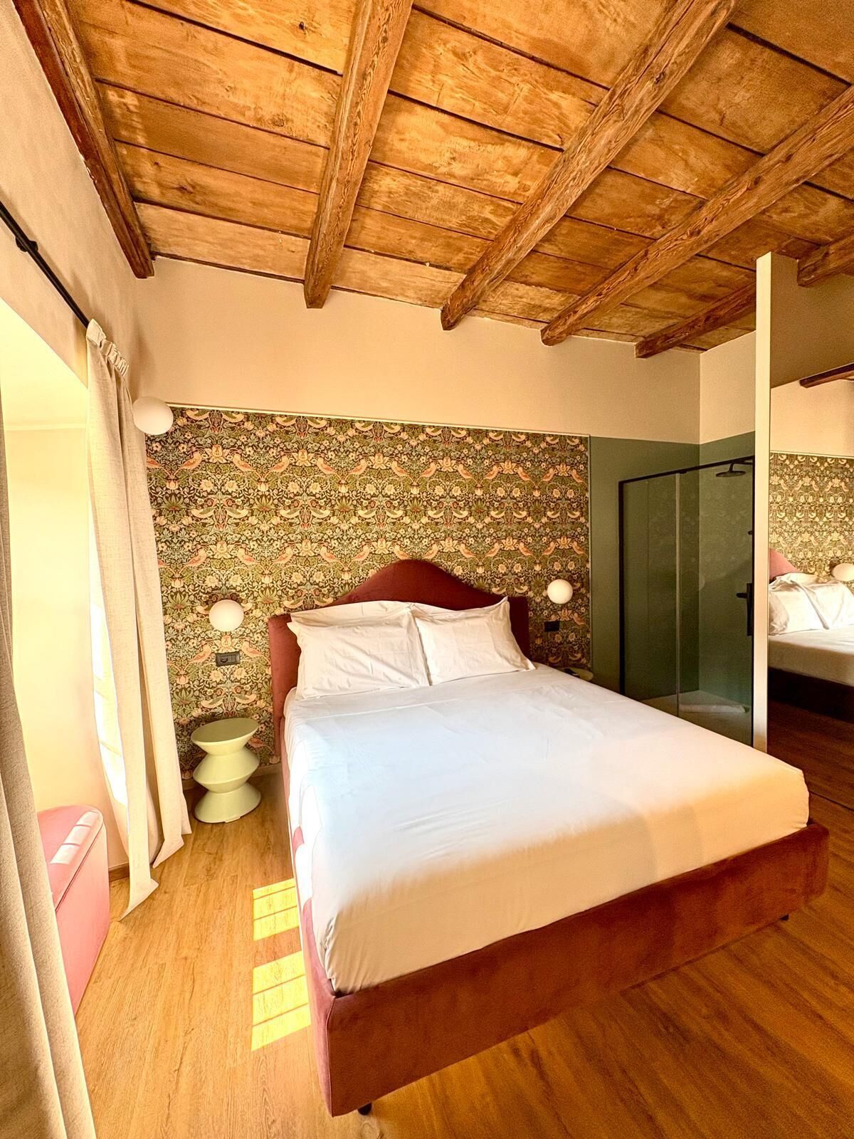 Un dormitorio con techo de madera, cama con ropa de cama blanca, pared con estampado, cabina de ducha de vidrio y una mesita de noche con lámpara.