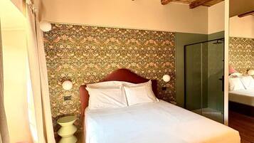 Un dormitorio con techo de madera, cama con ropa de cama blanca, pared con estampado, cabina de ducha de vidrio y una mesita de noche con lámpara.