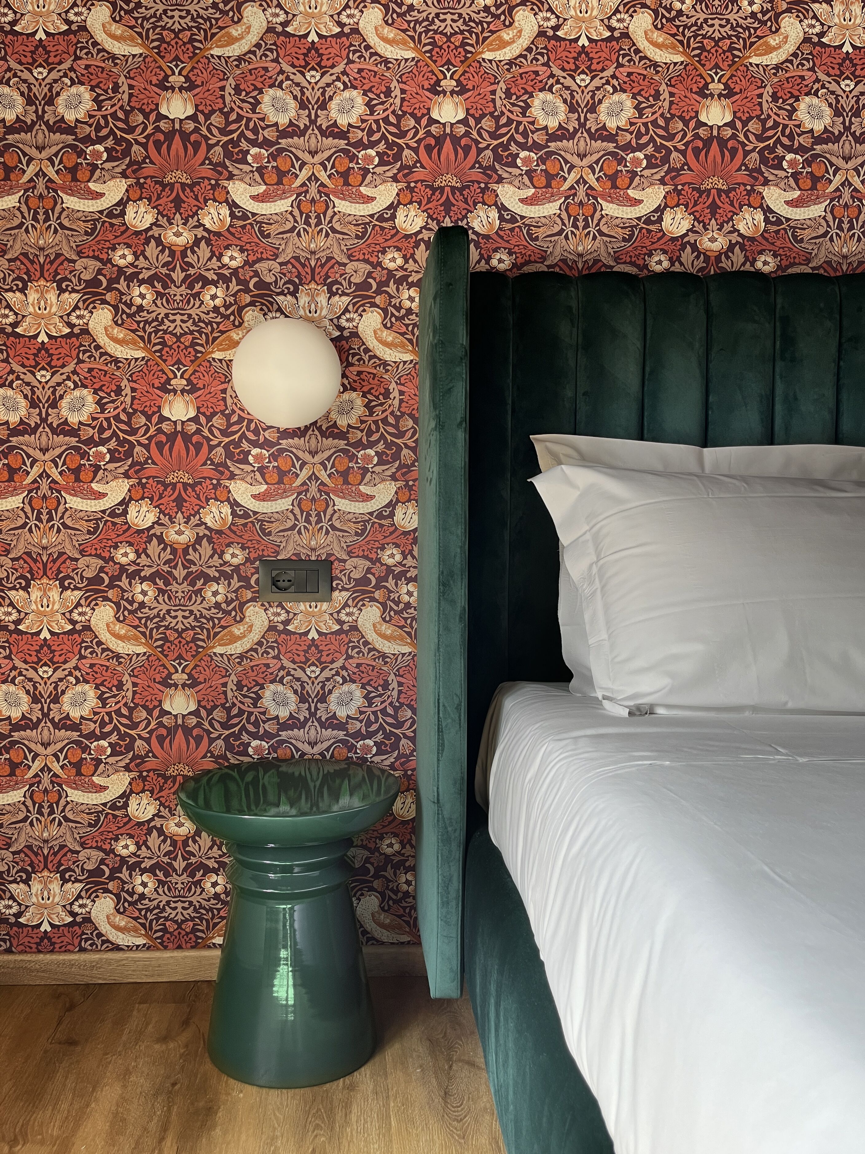 A room with a patterned wallpaper, a green bowl, a bed with white linens, and a dark green headboard.