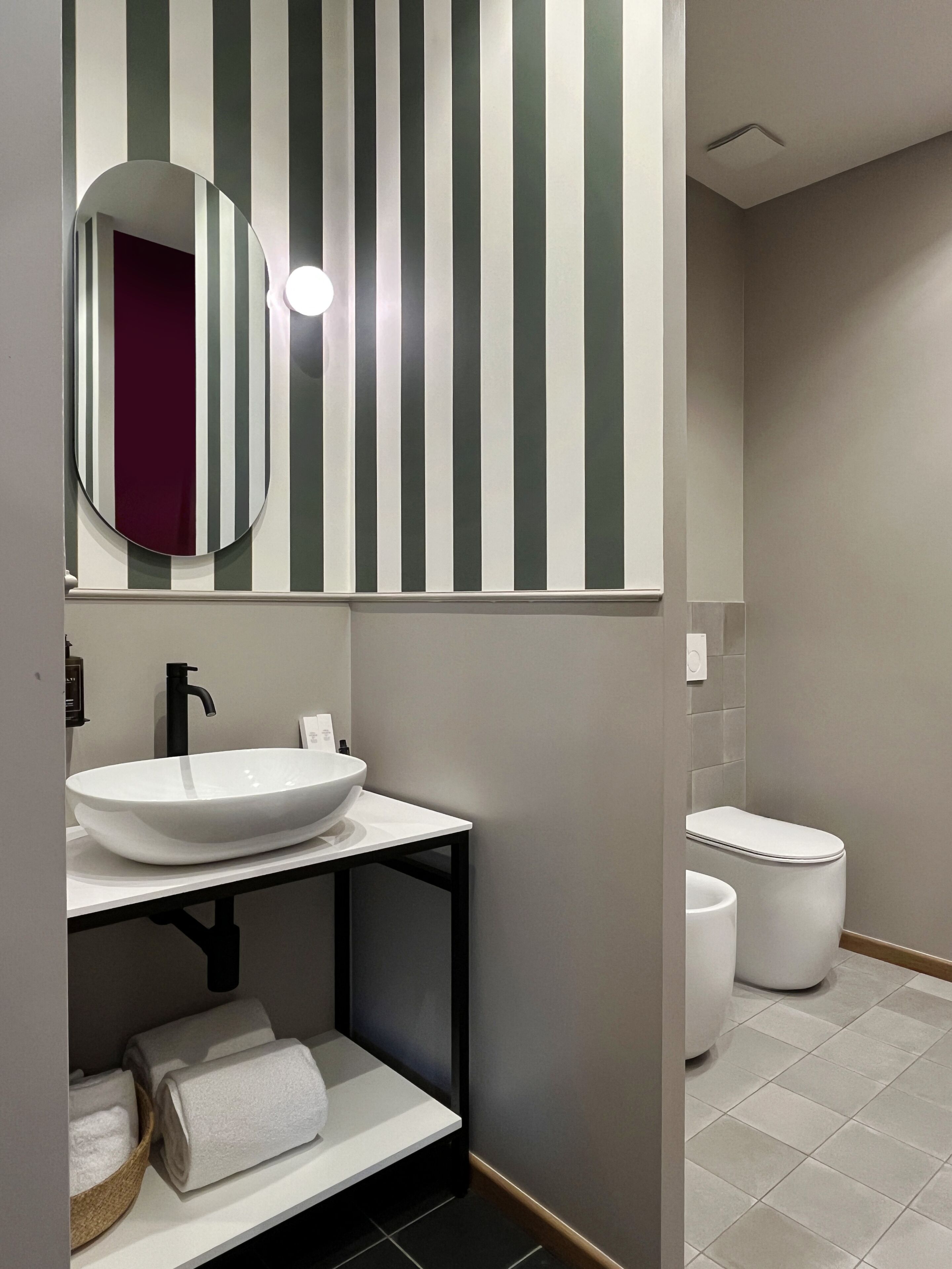 A modern bathroom with a white sink, black faucet, and a mirror with a round frame. There is a toilet and a towel rack with towels.