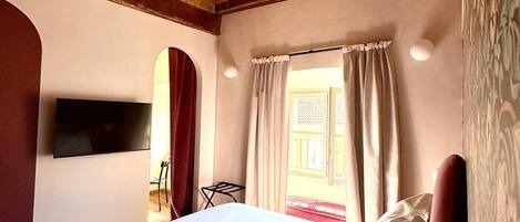 A room with a wooden ceiling, a bed with white linens, a TV mounted on the wall, and a window with curtains.