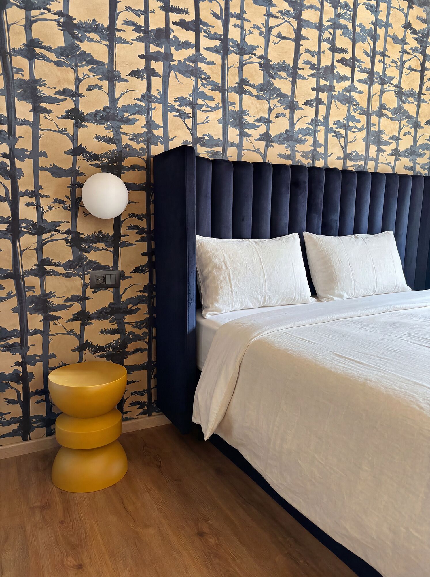 Quarto com cama, luminária na parede e um banquinho amarelo.