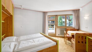 A room with a bed, a chair, a table, and a window with a view of buildings and trees. - Apartment 'Residence Ciasa Ulli Typ A' (San Cassiano)