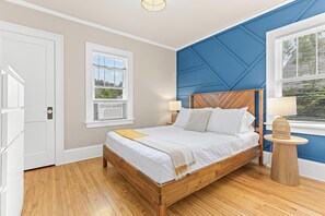 Une chambre à coucher avec un lit en bois, une literie blanche, une petite table ronde avec une lampe et une fenêtre avec des stores.
