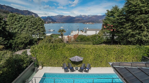 Una piscina con sillones de descanso y una sombrilla, rodeada de vegetación y con vistas a un lago con barcos.
