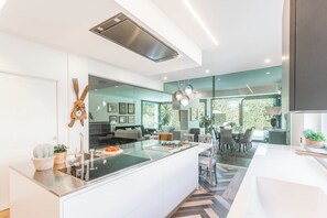 A modern kitchen with a central island, a dining area with chairs, and a view of greenery outside. - Villa Chiara - Four Bedroom Villa, Sleeps 8 (Stresa)