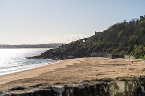 A beach with a rocky cliff, a clear sky, and a distant view of a town. - Hidden 19- Coastal Hot Tub (St Ives)