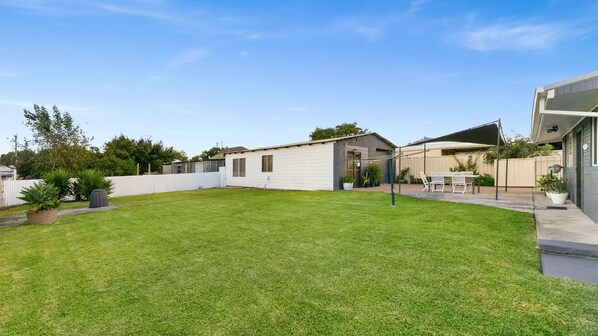 A residential backyard with a lawn, a patio area with a table and chairs, and a small shed.