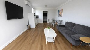 A modern living room with a grey sofa, a white coffee table with a plant, a flat-screen TV mounted on the wall, and a kitchen area with a refrigerator and a dining table.