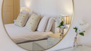 A bedroom with a bed, pillows, bedside lamps, a glass table, and a mirror reflecting the room.