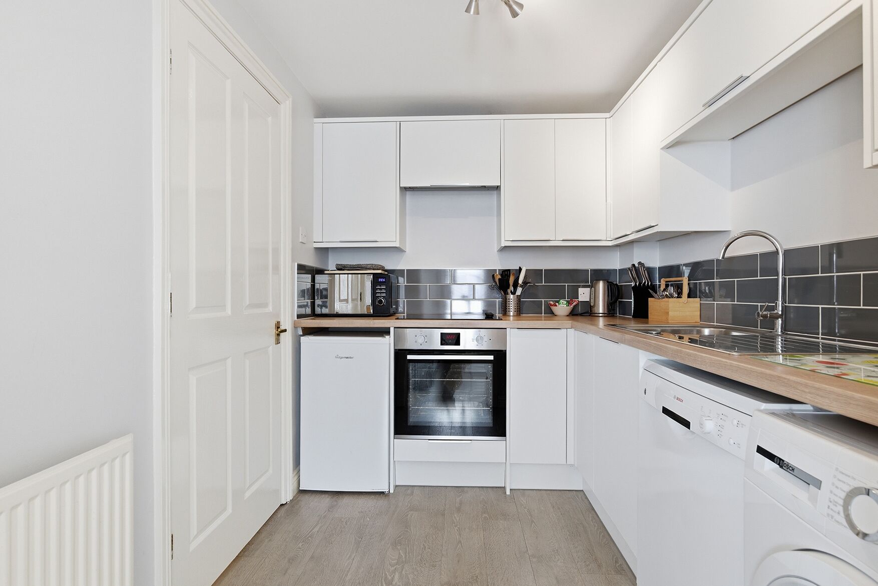 A compact kitchen with white cabinetry, a built-in oven, a dishwasher, and a countertop with a toaster and coffee maker.