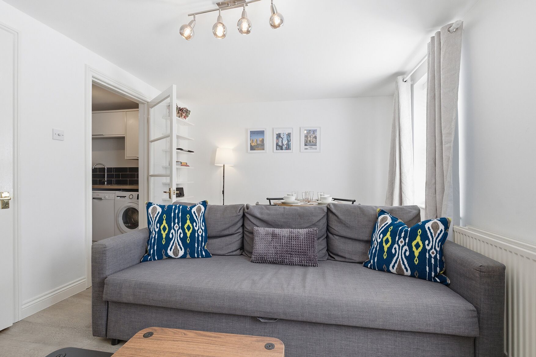 A living room with a grey sofa, patterned cushions, a coffee table, and a kitchen in the background.