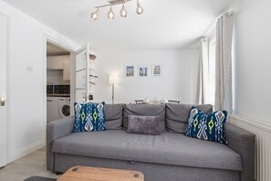 Una sala de estar con un sofá gris, cojines estampados, una mesa de centro y una cocina al fondo.