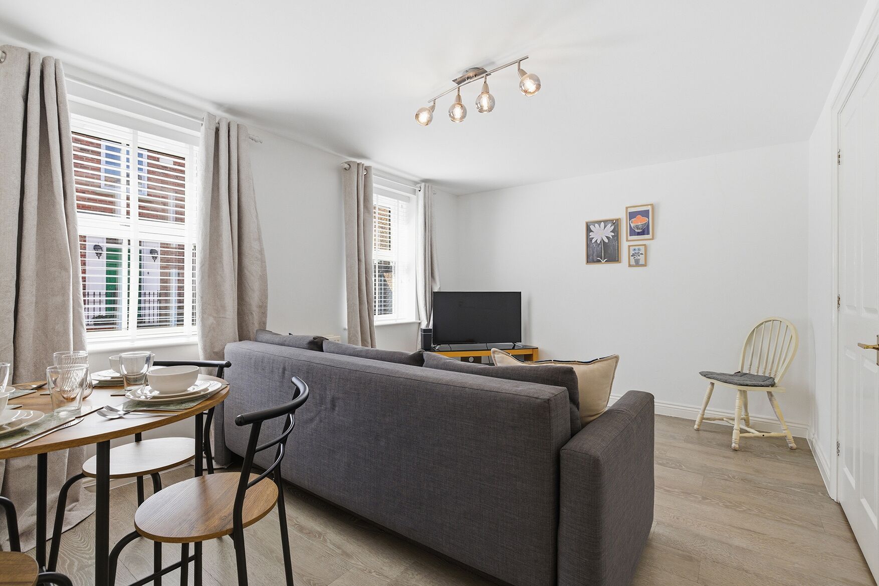 A modern living room with a grey sofa, a dining table with chairs, a television, and a wooden floor.