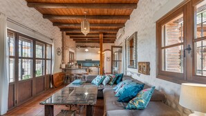Une pièce spacieuse avec un plafond en bois, de grandes fenêtres, un canapé agrémenté de coussins colorés, une table en verre et un panier en osier.