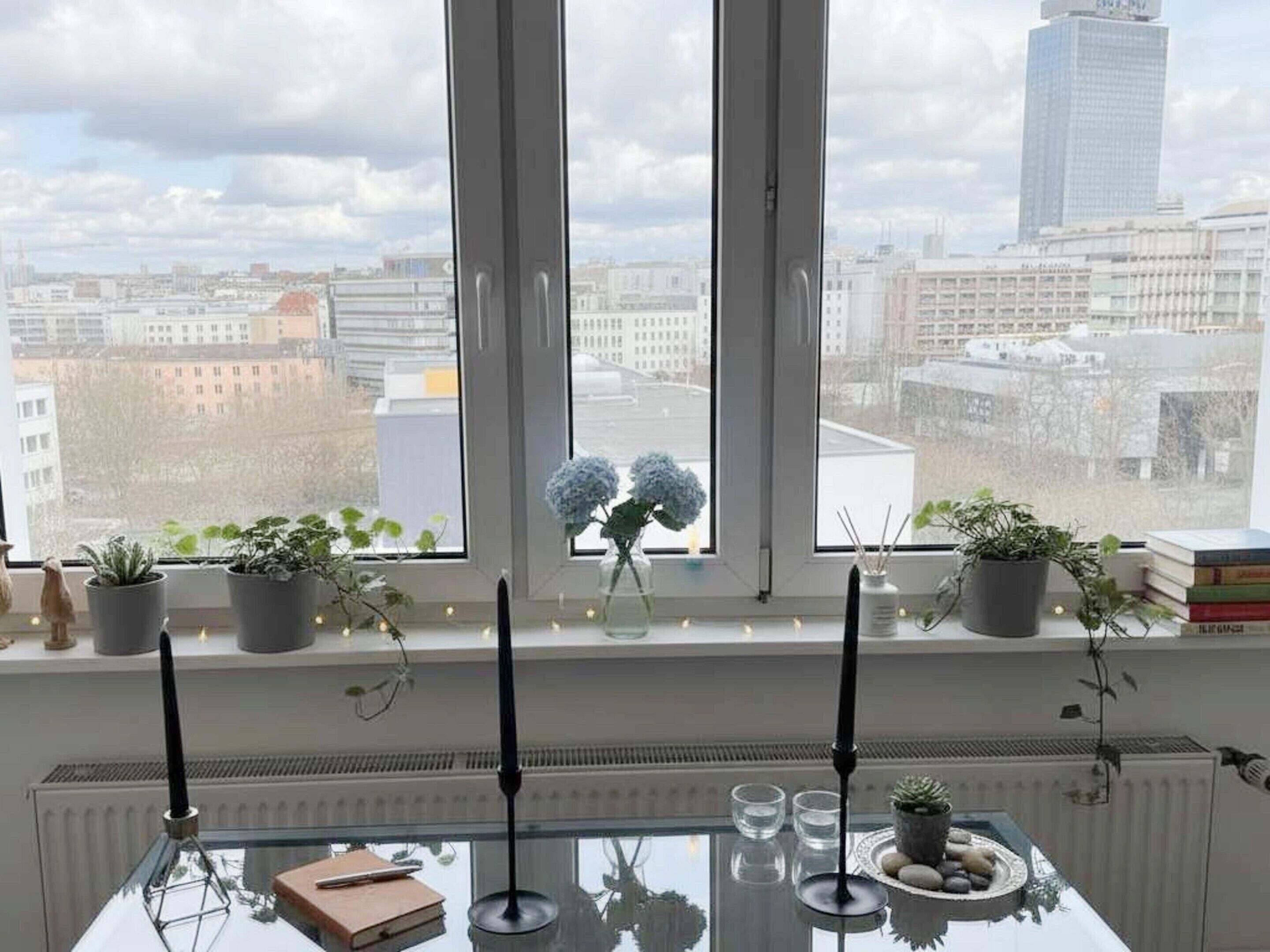 A room with a view of a cityscape through large windows, a table with a book and candles, and potted plants on the windowsill.