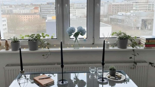 A room with a view of a cityscape through large windows, a table with a book and candles, and potted plants on the windowsill. - Sunny Stay in Alexanderplatz (Berlin)