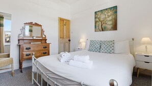 Ein Schlafzimmer mit einem Bett, einer hölzernen Kommode, einem Spiegel, einer Tür, einem Gemälde und zwei Nachttischen mit Lampen.