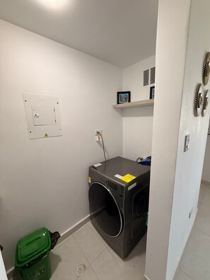 Ein Waschraum mit einer Waschmaschine, einem grünen Müllbehälter, einem Regal mit einem Bilderrahmen und einem an der Wand befestigten Stromverteiler.