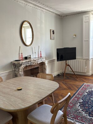 Una habitación con chimenea de mármol, una mesa redonda con sillas, un televisor sobre un trípode y un tapete estampado.