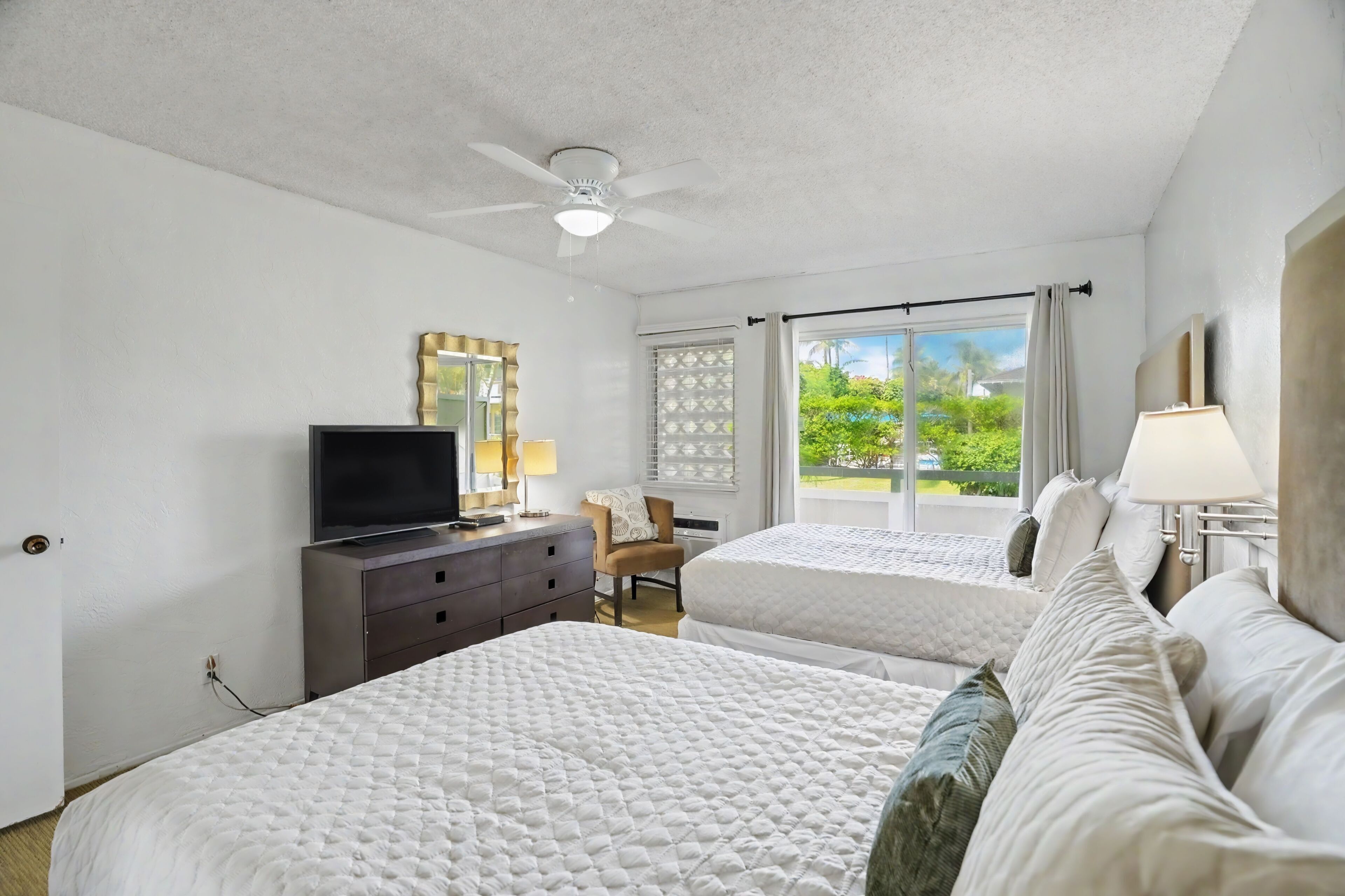 A hotel room with two beds, a TV, a ceiling fan, a desk, and a view of greenery outside.
