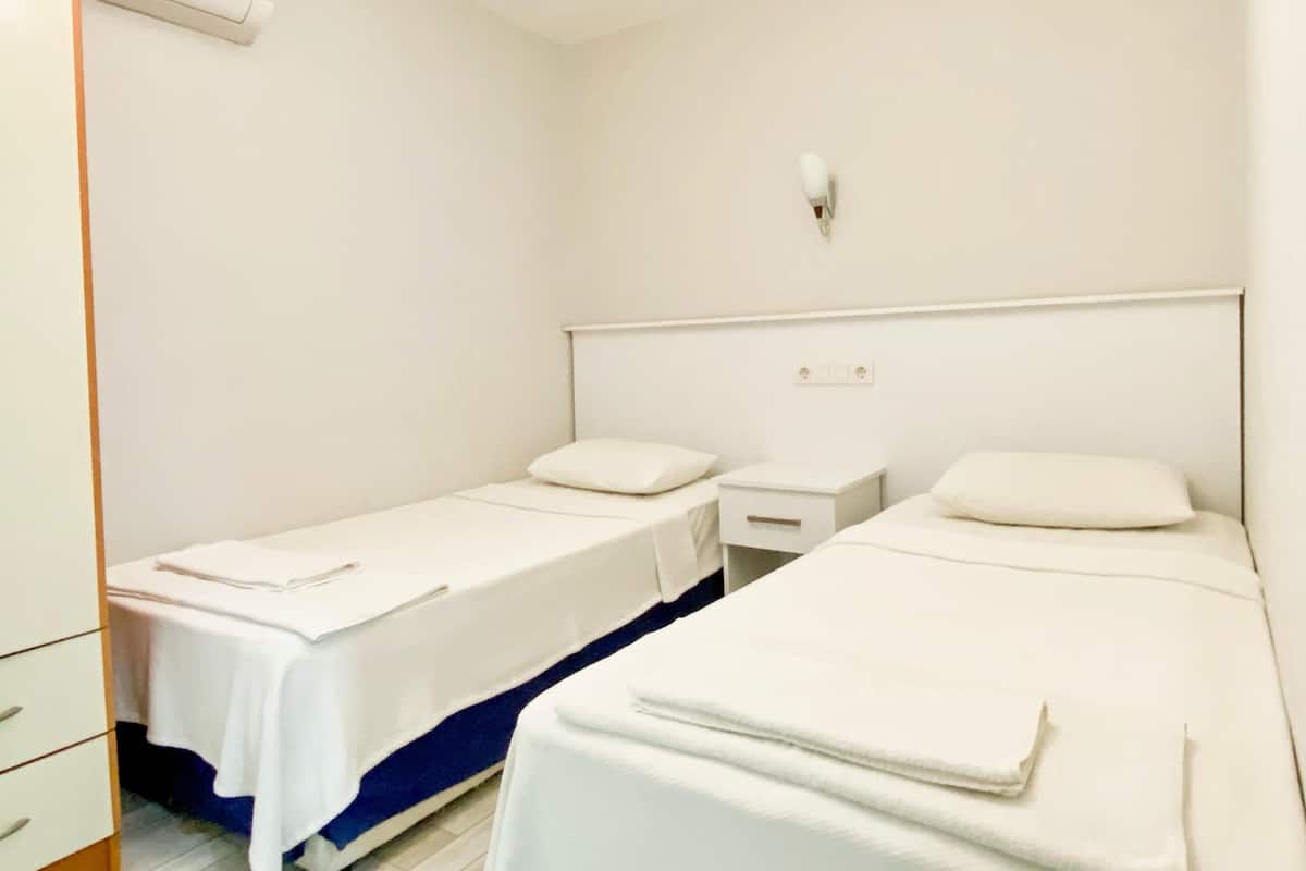 Habitación de hotel con dos camas individuales, una mesita de noche con lámpara, un armario y un equipo de aire acondicionado.