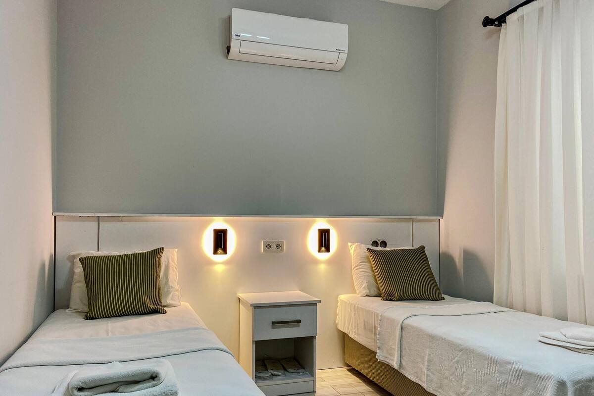 Habitación de hotel con dos camas, una mesita de noche, aire acondicionado e iluminación en la pared.
