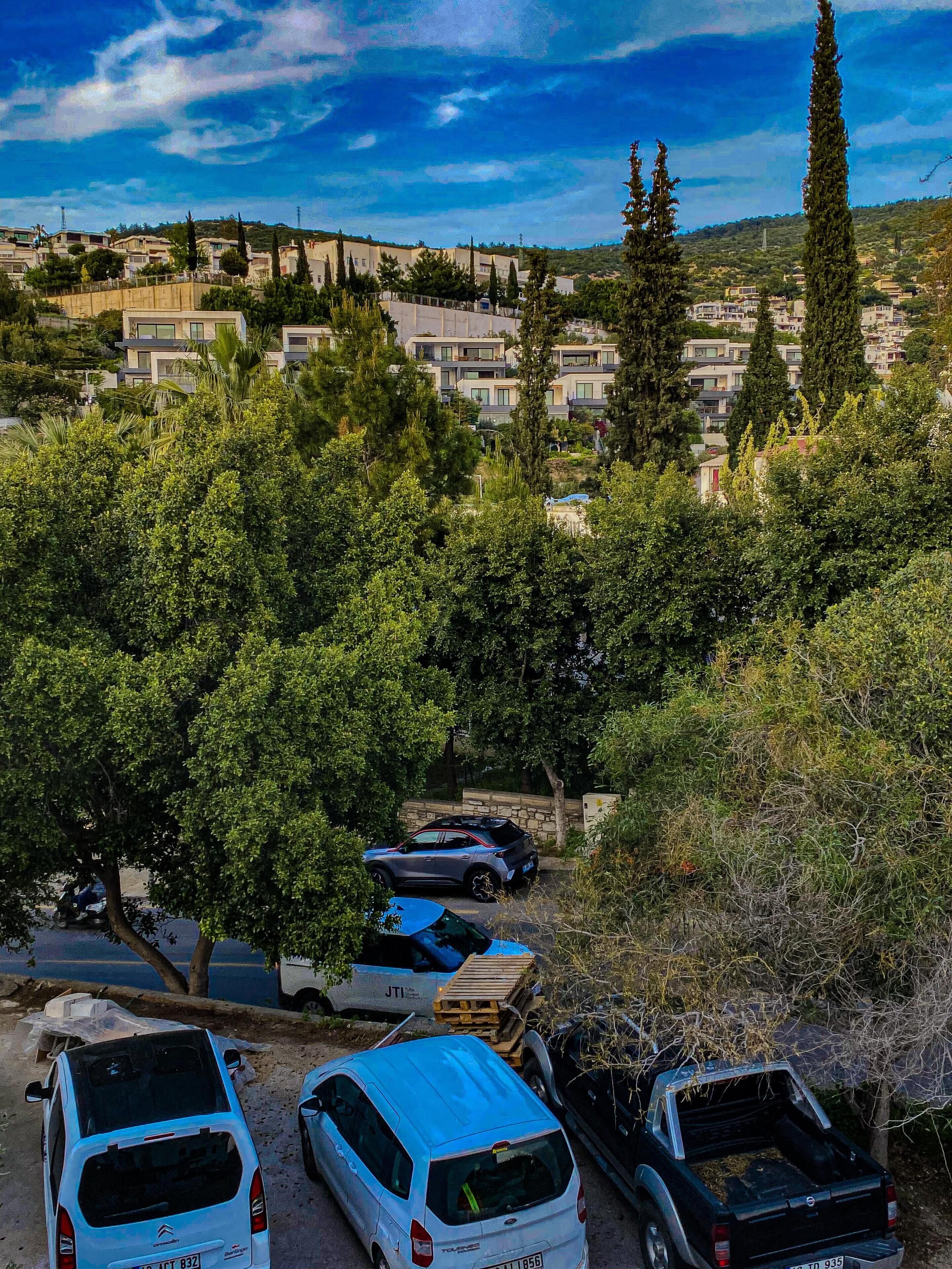 Ein Parkplatz mit mehreren Autos, umgeben von Bäumen und Gebäuden im Hintergrund.