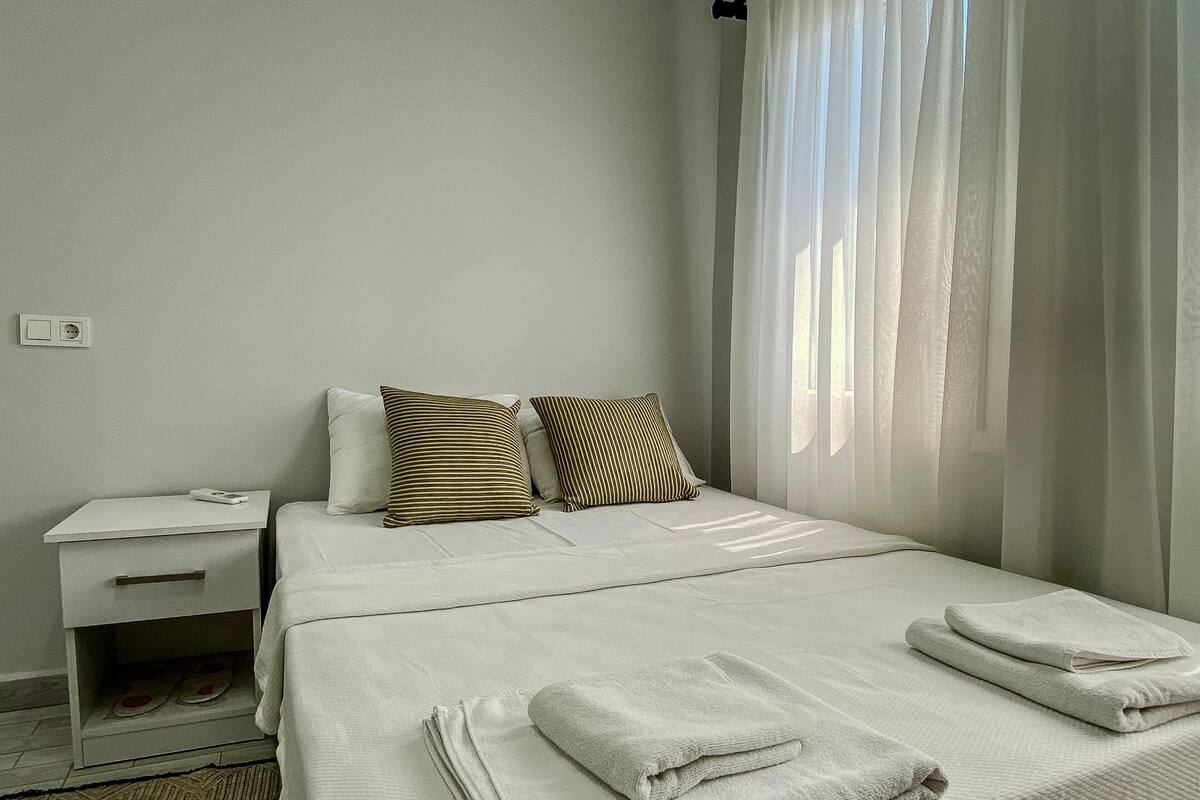 Una habitación de hotel con cama, almohadas, mesita de noche y ventana con cortinas.