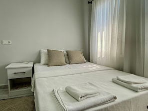 Una habitación de hotel con cama, almohadas, mesita de noche y ventana con cortinas.