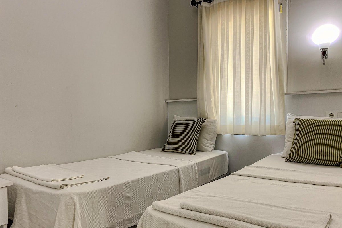 Una habitación con dos camas individuales, una mesita pequeña, una lámpara de pared y un ventanal con cortinas traslúcidas.