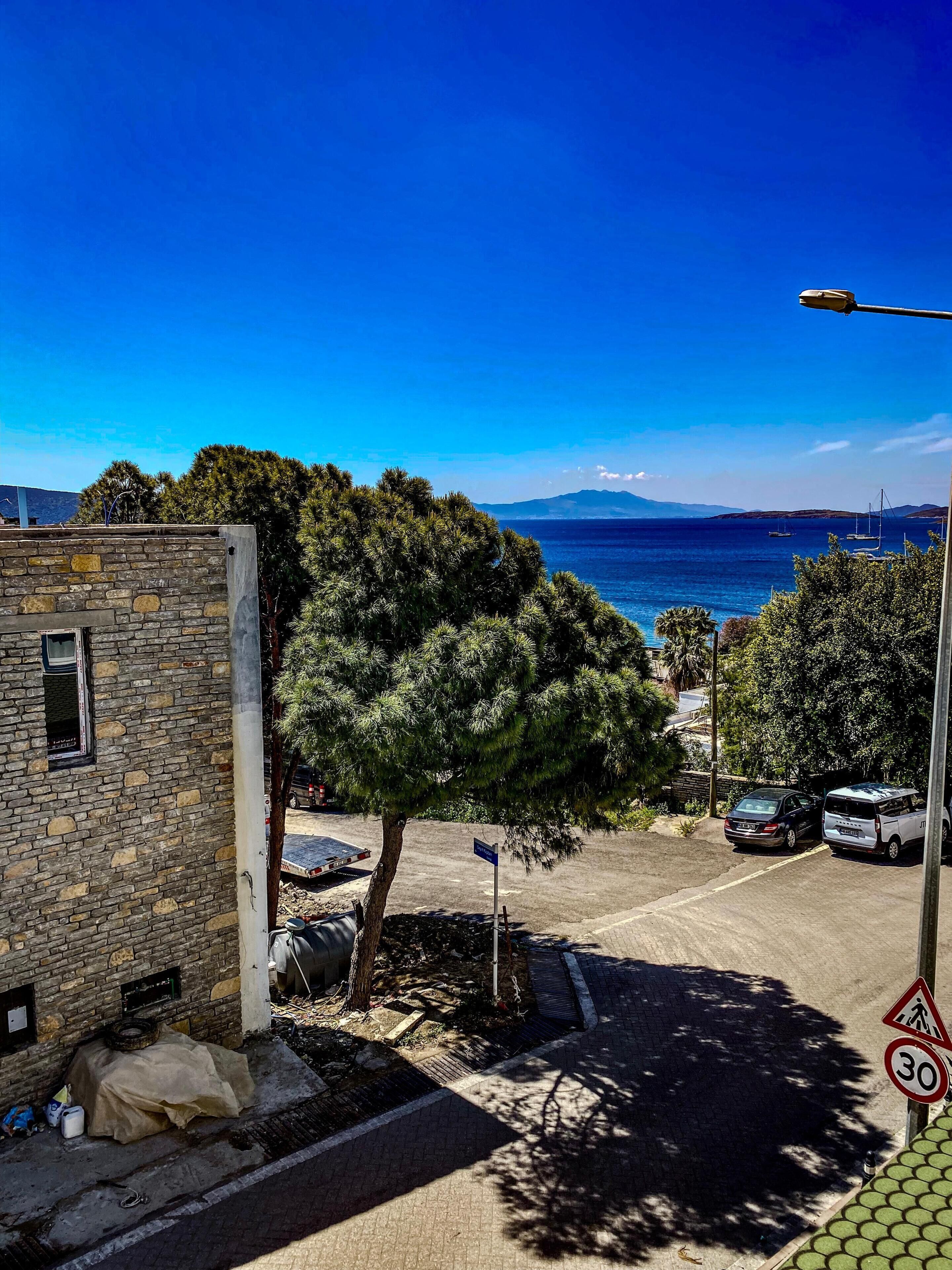 A coastal street with parked cars, a stone building, and a clear view of the sea and mountains in the distance.