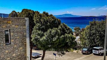A coastal street with parked cars, a stone building, and a clear view of the sea and mountains in the distance.